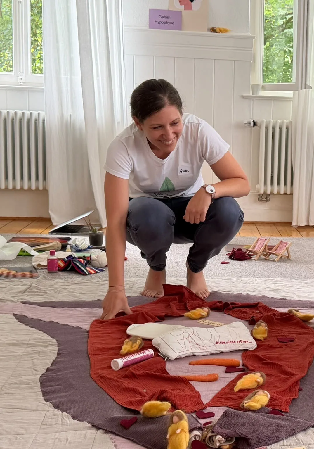Anne hockt lächelnd neben dem großen Bodenbild, auf dem Tücher, Filzfiguren und Alltagsgegenstände den Zyklus darstellen
